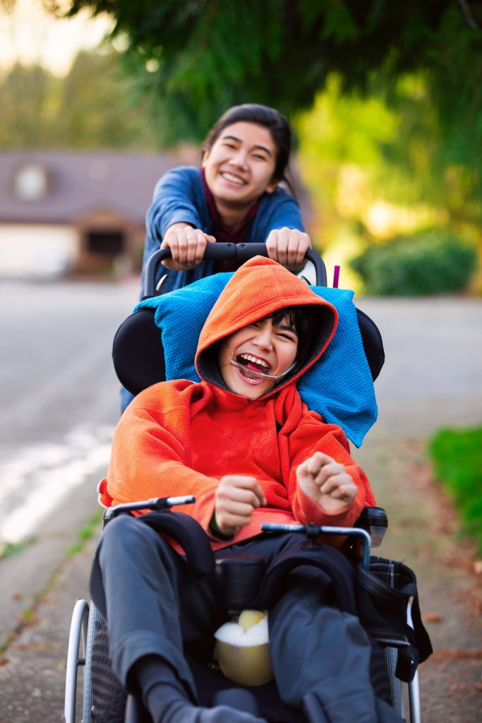 Big sister pushing little brother in wheelchair around neighbourhood, laughing and smiling